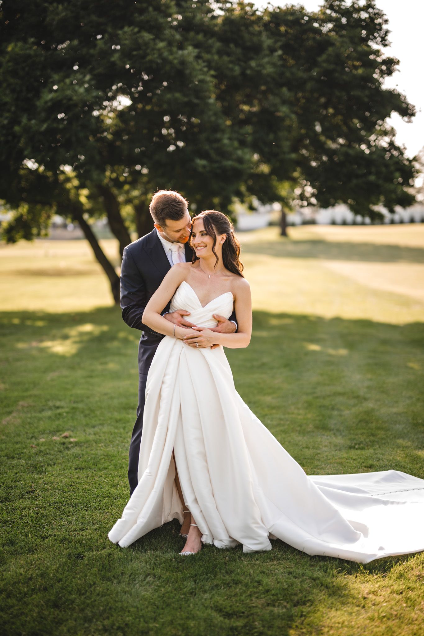 Elegant wedding couple poses on manicured lawn with flowing white dress.
