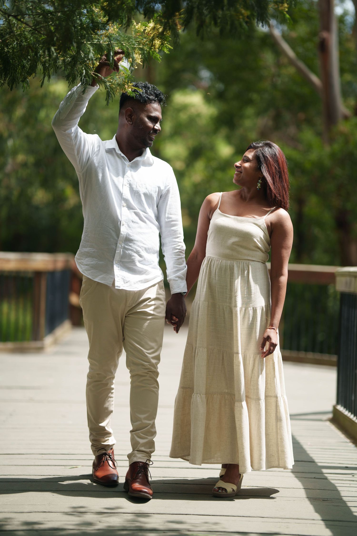A couple in white outfits walking together on a sunny wooden boardwalk through a park.