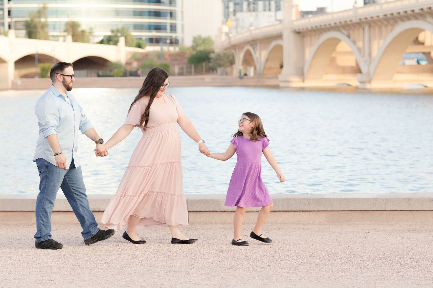 Family of 3 holding hands and walking in a line with the young daughter leading and looking back at parents in front of Tempe Town Lake.