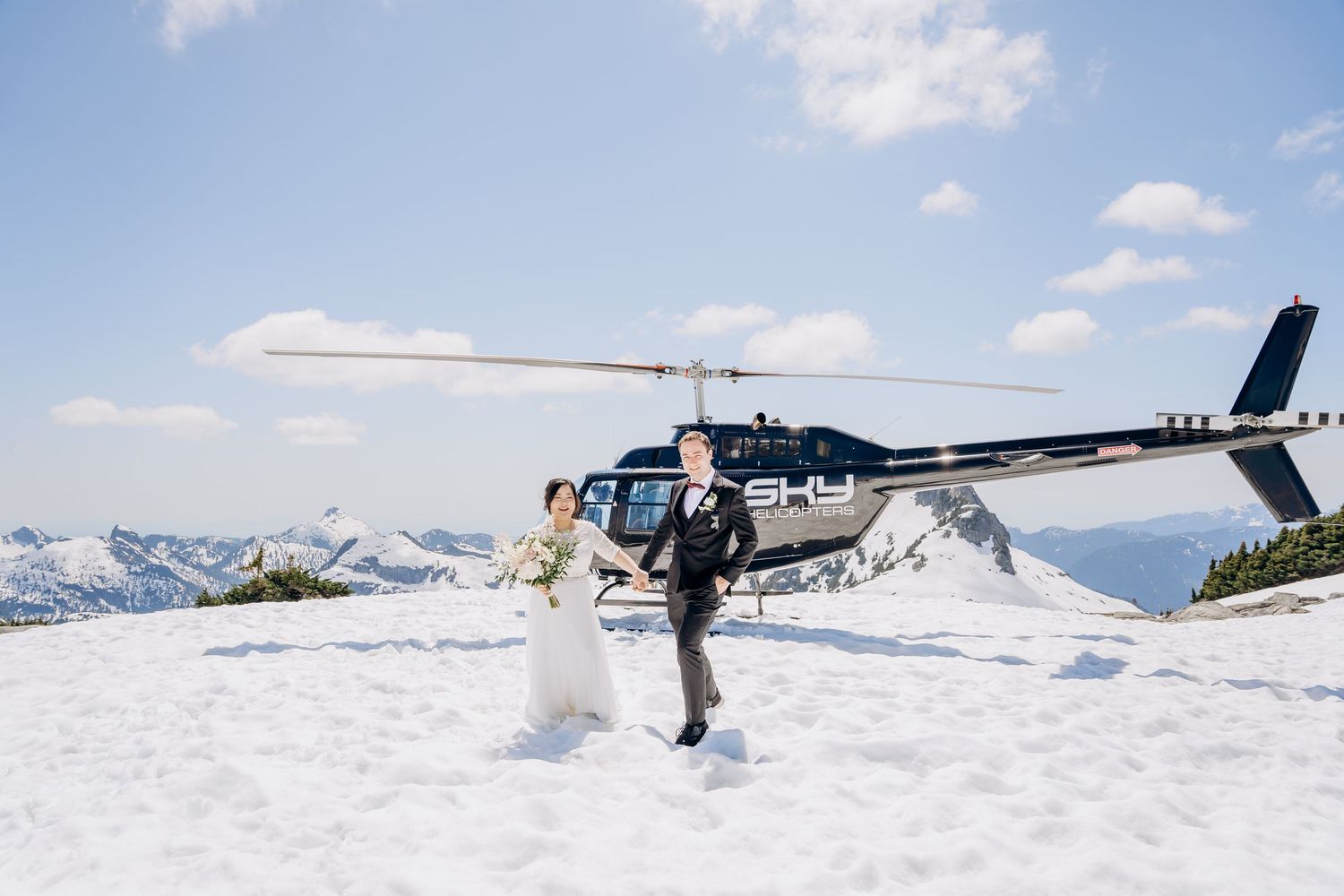 A wedding couple poses in front of a helicopter on a snowy mountain peak.