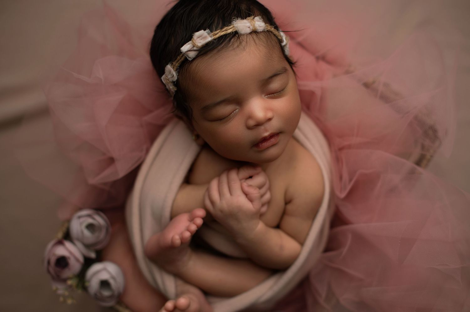Sleeping newborn in pink tulle with silver headband rests peacefully in cozy beige wrap during professional photo session.