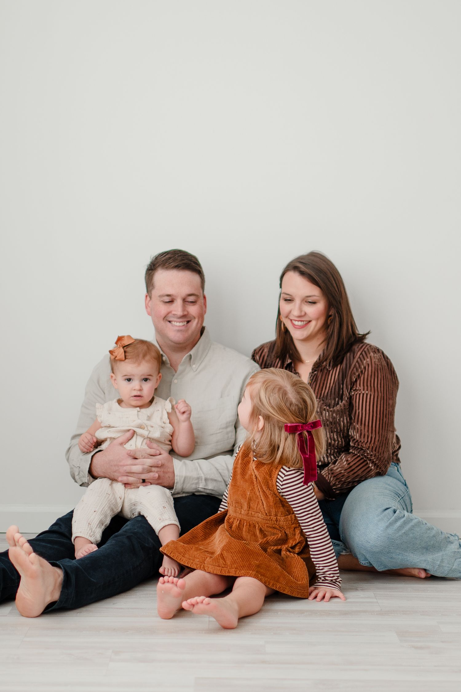 Family sits together on light wooden floor against white wall for casual indoor portrait session.