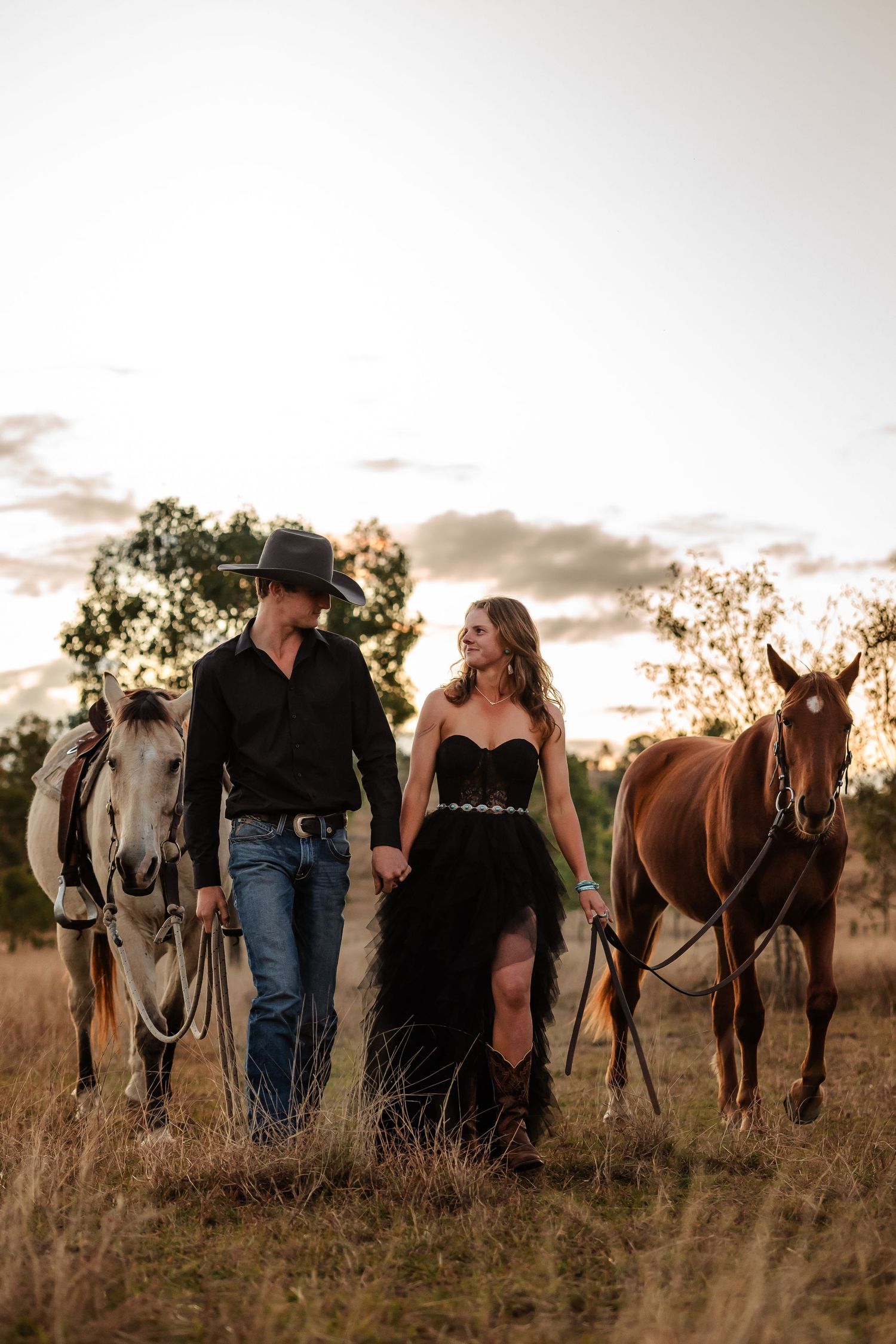 Couple stands with their horses in tall grass field during golden sunset, creating a romantic silhouette.