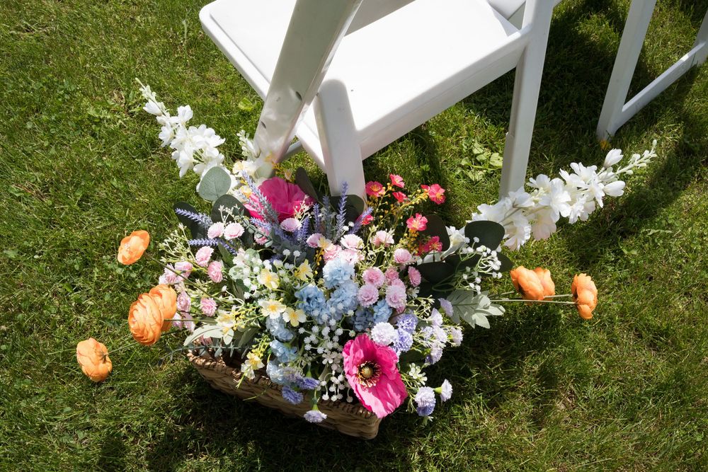 Colorful floral arrangement with peach roses, pink blooms and white delphinium on grass next to a white garden chair.
