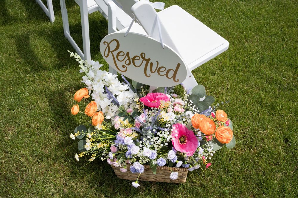 A white chair with colorful floral arrangements featuring pink and purple flowers sits on lush green grass in a garden setting.