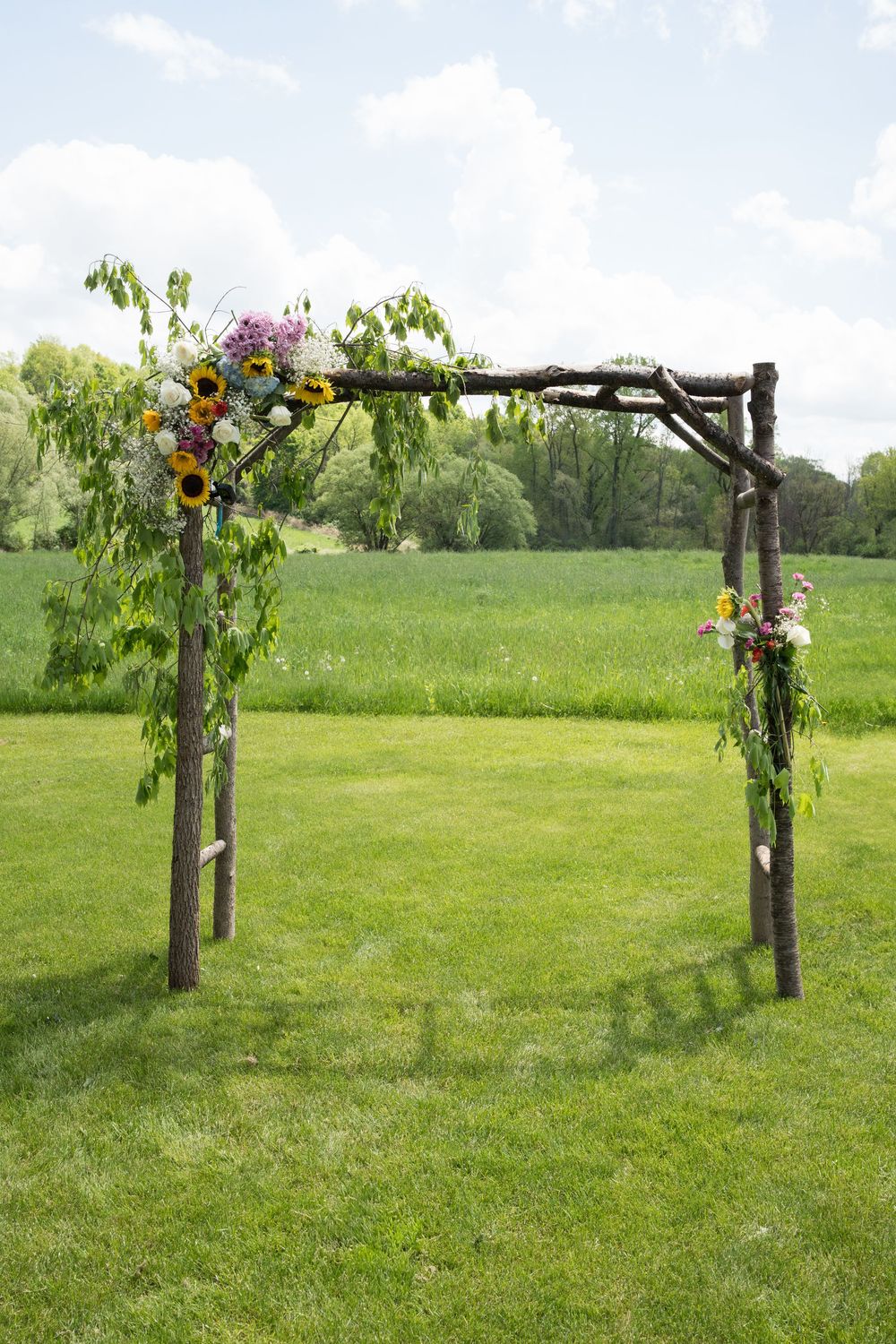 Decorative wooden posts with floral arrangements form an outdoor wedding arch pathway across a lush green lawn.