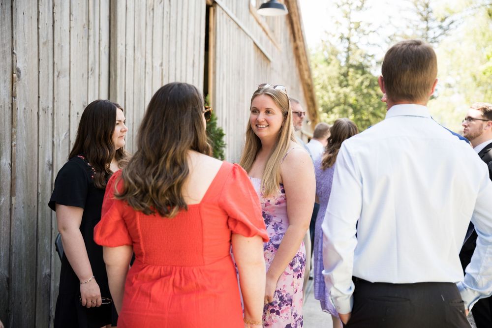 Outdoor wedding ceremony guests gathered by rustic wooden barn walls on a sunny afternoon.