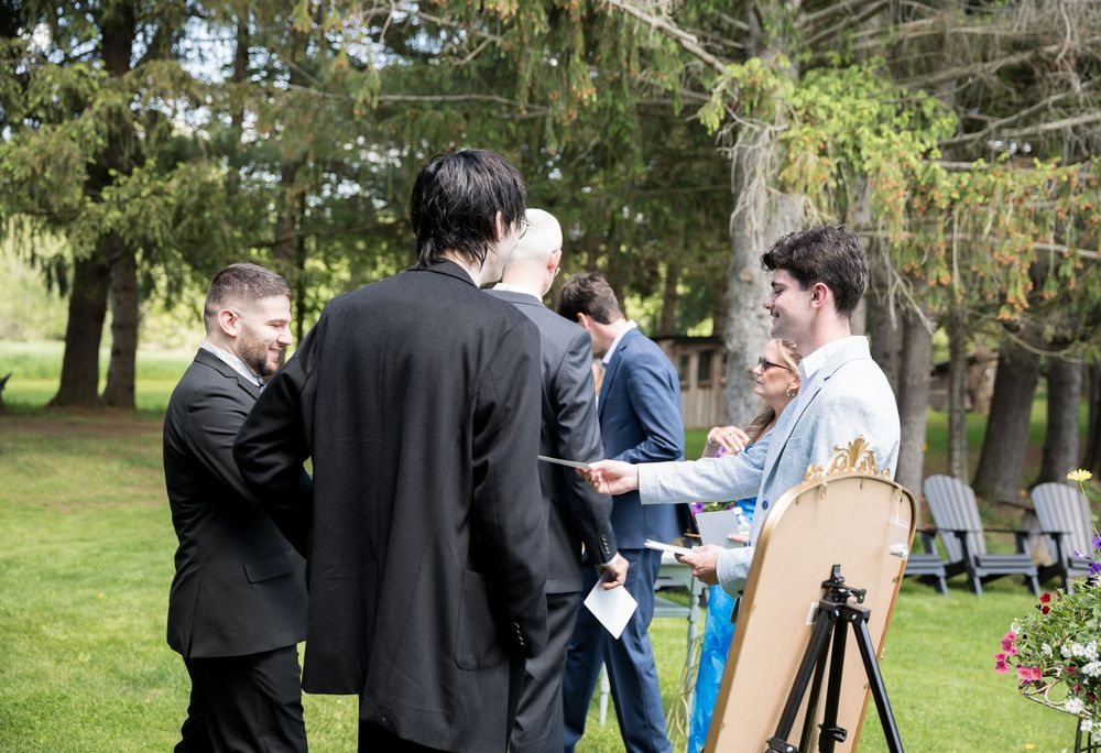 A group of formally dressed people gathered for an outdoor social event in a garden or park setting with trees.