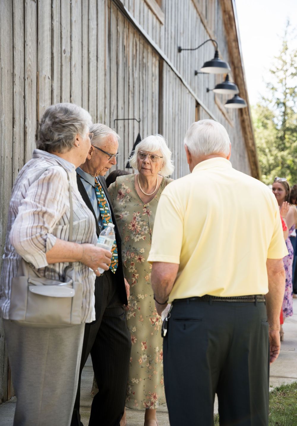 Family members help and embrace elderly person leaning on support pillar at outdoor gathering venue.