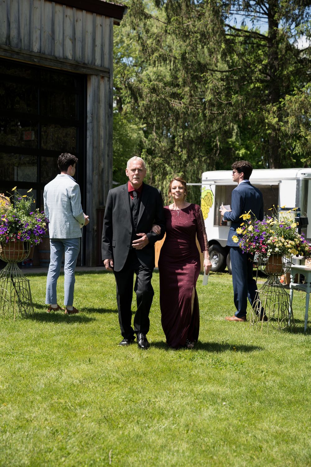 People socialize on a sunny lawn beside tall trees at an outdoor gathering.
