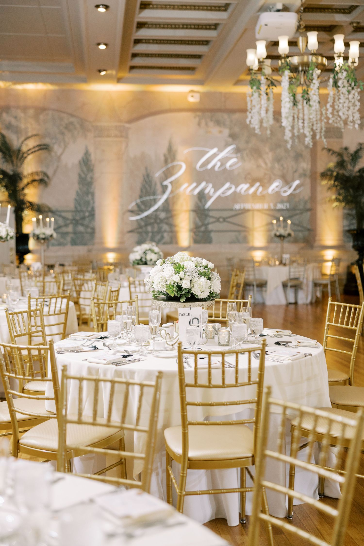 Elegant wedding reception ballroom with crystal chandelier, gold chiavari chairs, and white tablescapes.