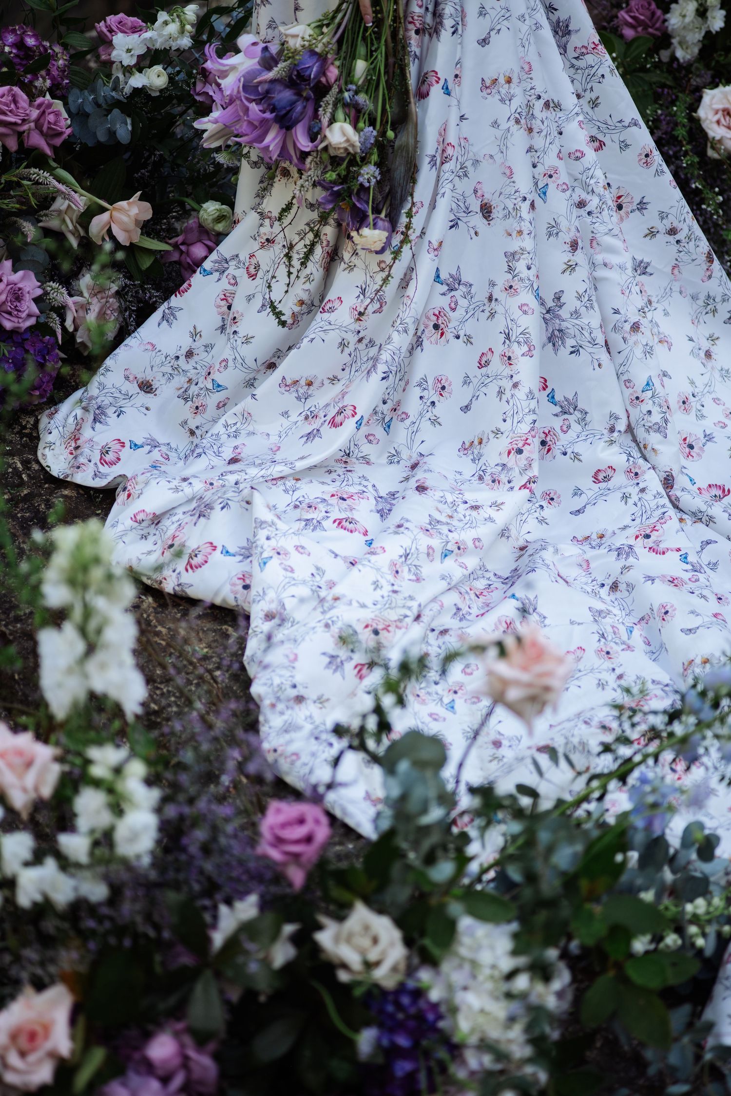 Flowing floral bridal gown train spreads among purple, white, and pink wedding flowers on ground.