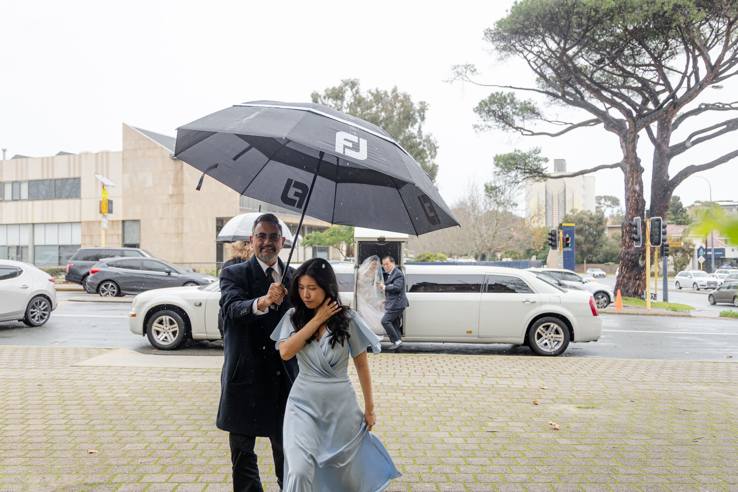 A couple shares an umbrella while standing together in a grey urban parking lot on an overcast day.