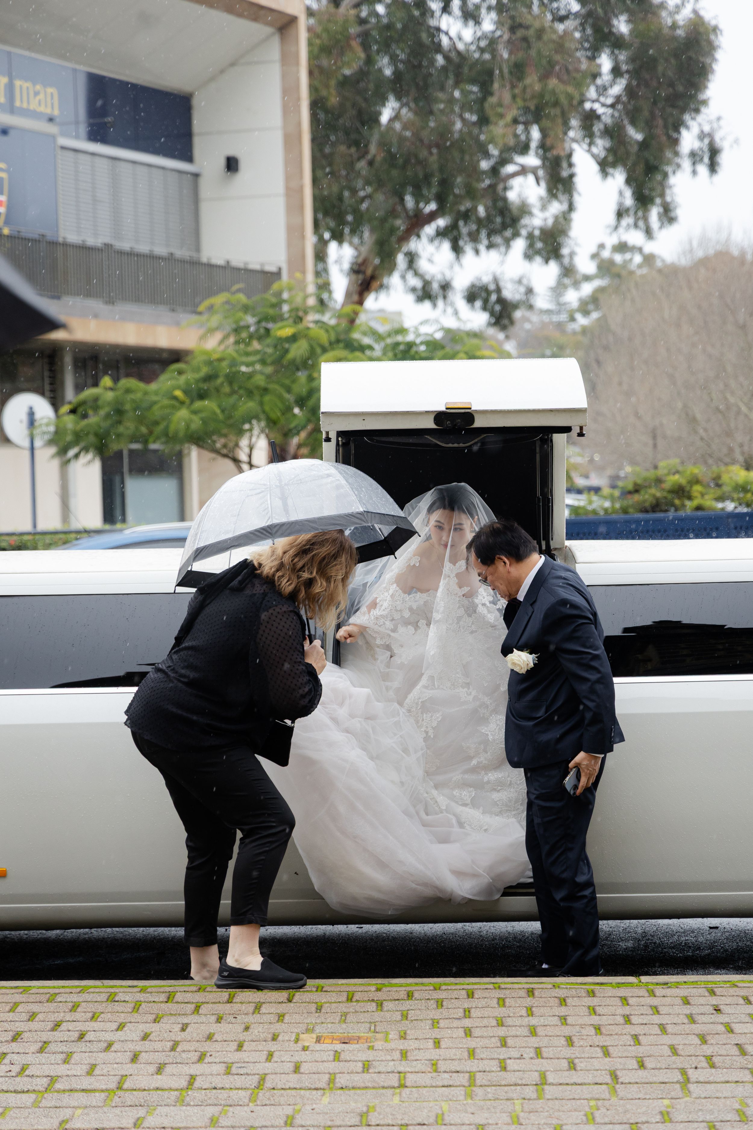 A bride in white dress being helped into a white stretch limousine on a rainy day.