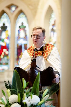Clergy member wearing ornate robes stands at altar with white lily floral arrangements.
