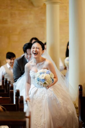 Joyful wedding scene with a bride in a white lace gown and veil laughing while holding a blue and white bouquet.