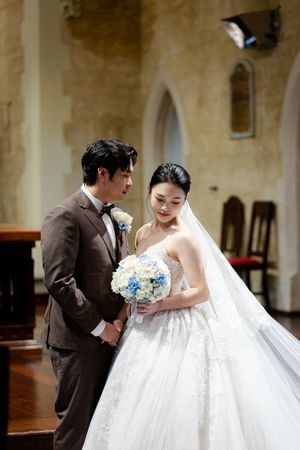 Couple in wedding attire posing in an elegant church setting with a blue and white bouquet.