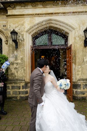 Newlyweds share a romantic kiss outside a historic stone church building as rice confetti rains down.