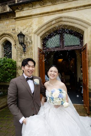 Newlyweds celebrate outside historic stone church building under falling confetti.