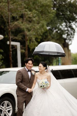 A wedding couple poses with black umbrellas next to a white luxury car on a tree-lined street.