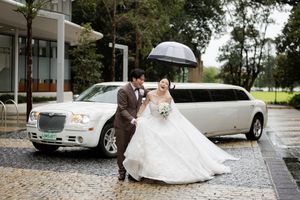 A couple on their wedding day standing next to a silver Chrysler limousine under umbrellas in the rain.