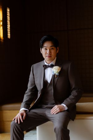 A formal portrait of someone in a dark brown tuxedo seated against a dramatic black background.