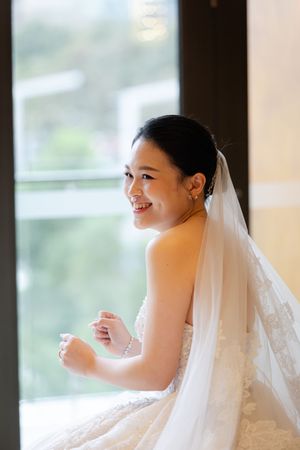 Bride in a flowing white dress and veil smiles while looking out a modern window.