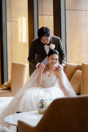 A tender moment between a bride in a white wedding gown and groom in a dark suit sharing an embrace on a beige couch.
