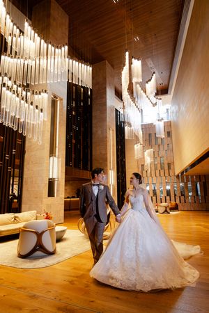 Elegant wedding couple in formal attire pose under dramatic crystal lighting in modern church space with wooden floors.