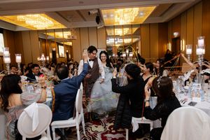 Wedding reception scene showing guests celebrating at decorated tables with crystal chandeliers overhead.