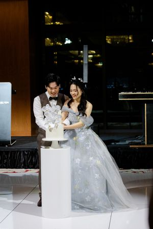 A couple at their wedding reception laughs together while cutting their white wedding cake on an elegant display stand.