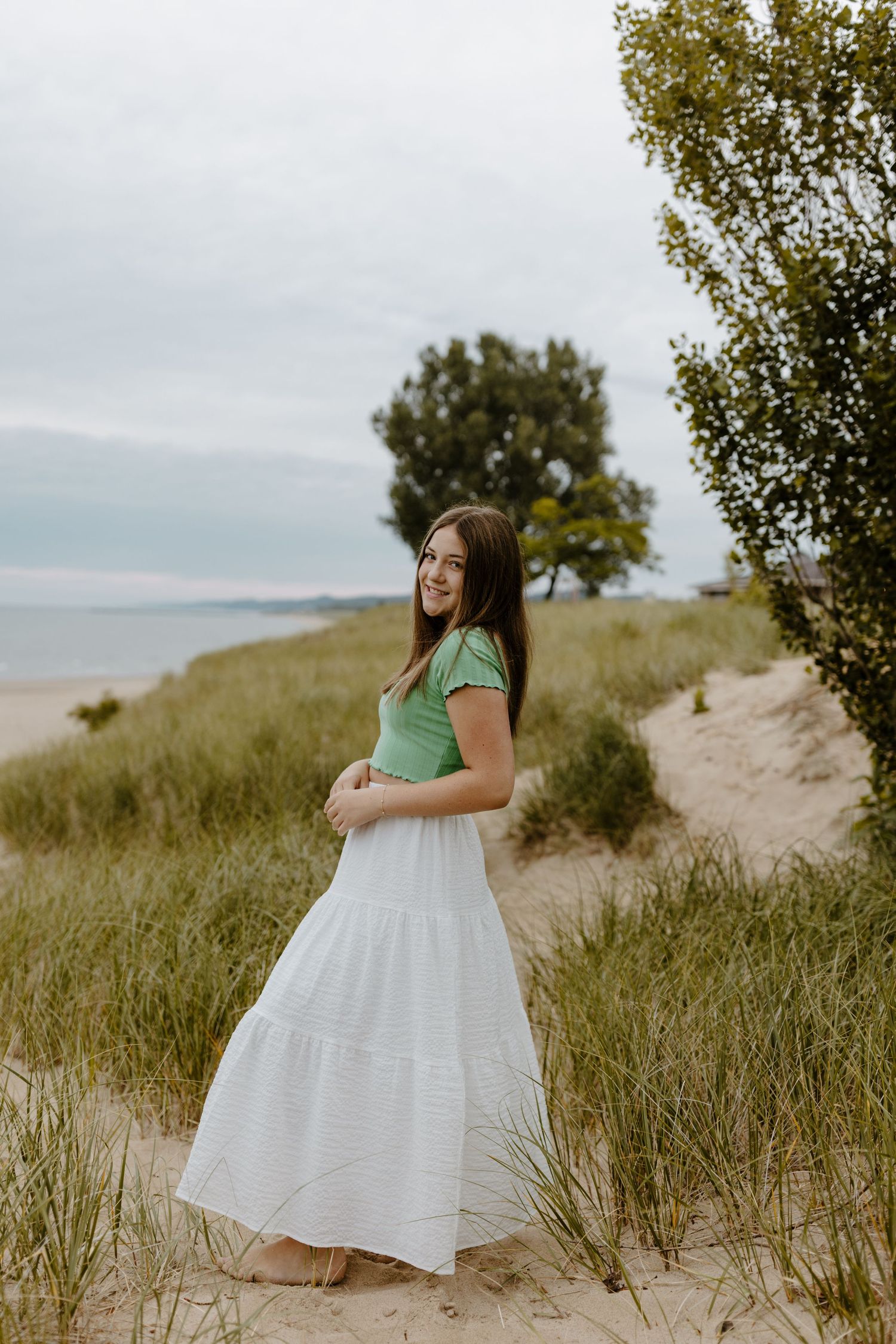 A young woman in a green top and white skirt stands in a field near a body of water and trees. Fort Worth Texas senior photographer.