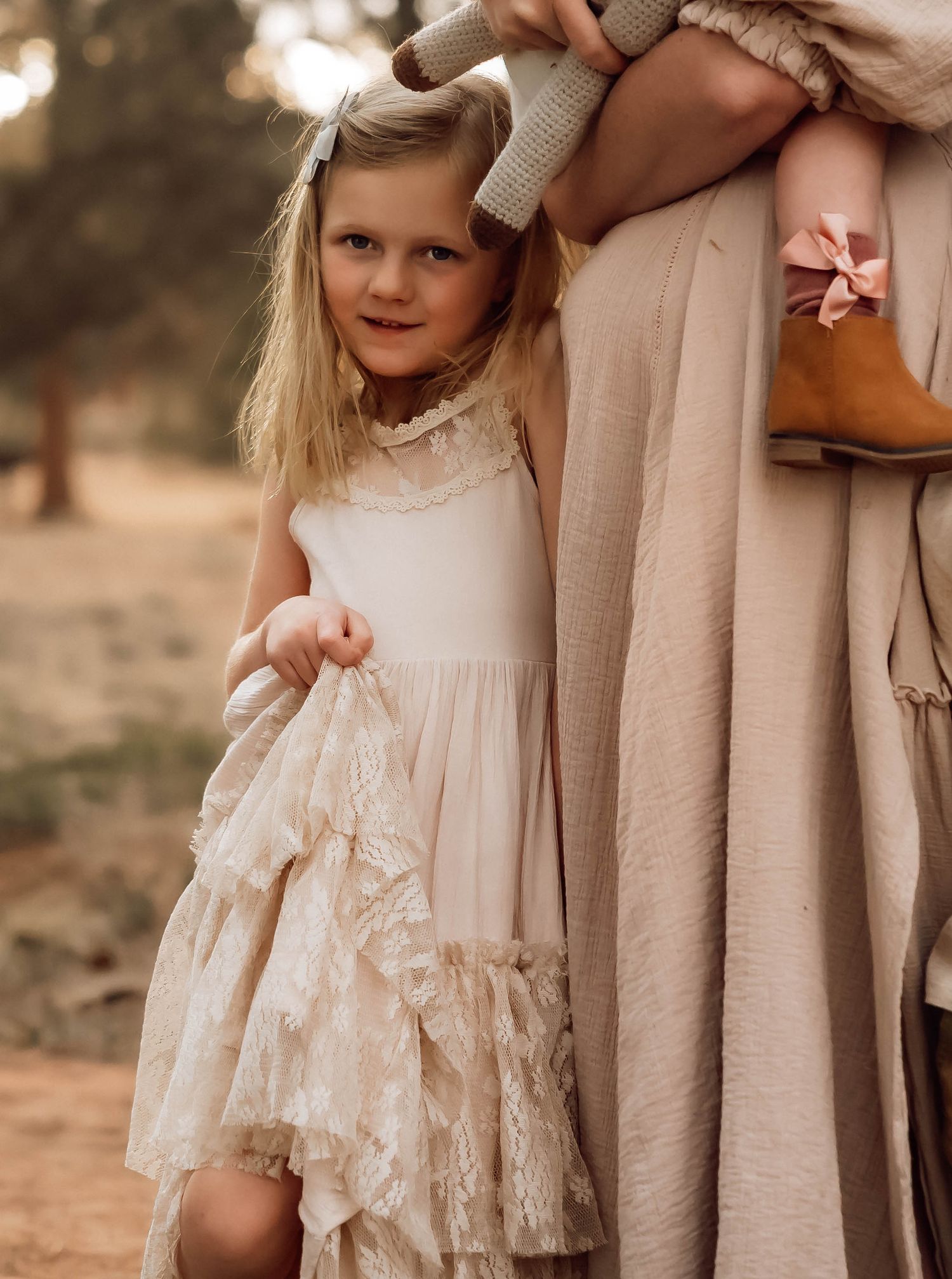 A young girl in a lace dress holds fabric while standing next to flowing neutral clothing.