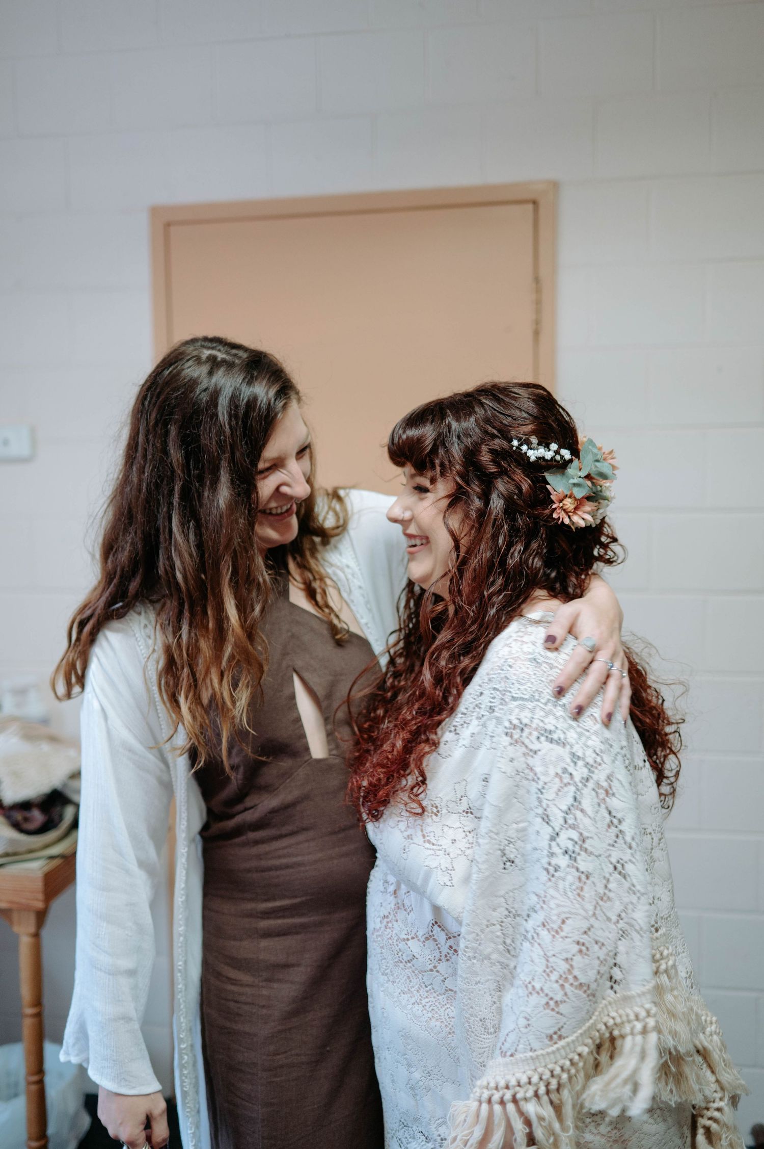 Two people sharing a joyful moment together, one wearing a brown dress and one in a bohemian style lace wedding dress.