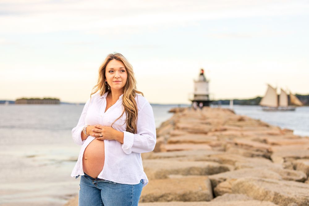 Expectant mother in white shirt and jeans stands on rocky jetty by lighthouse during golden hour.