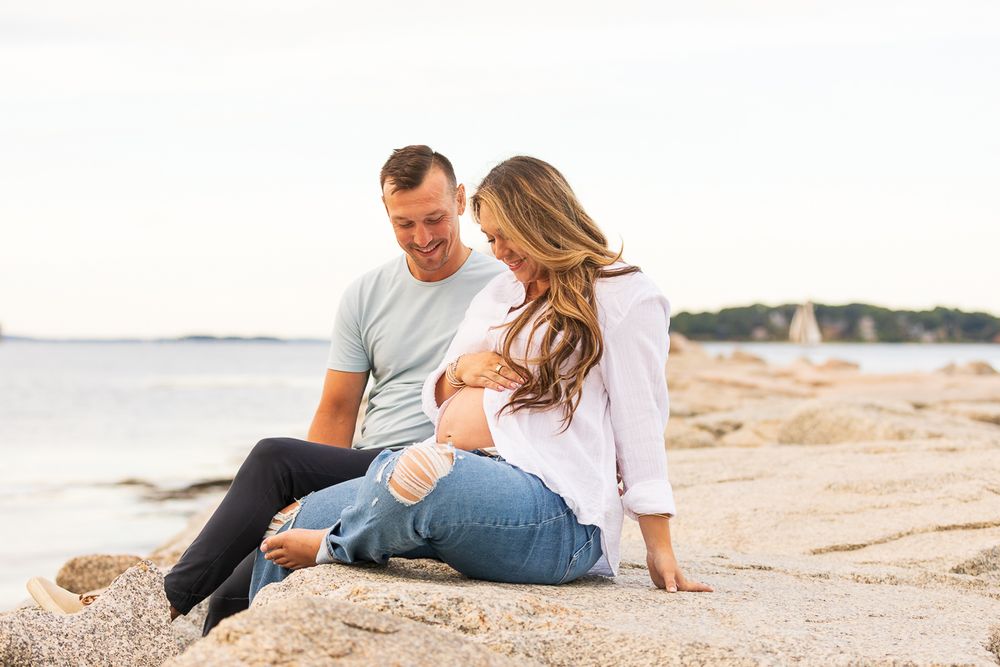 A couple sits together on a beach at sunset with sailboats visible in the distance.
