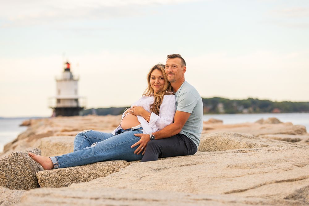 A couple sits together on coastal rocks at sunset with a lighthouse in the background.