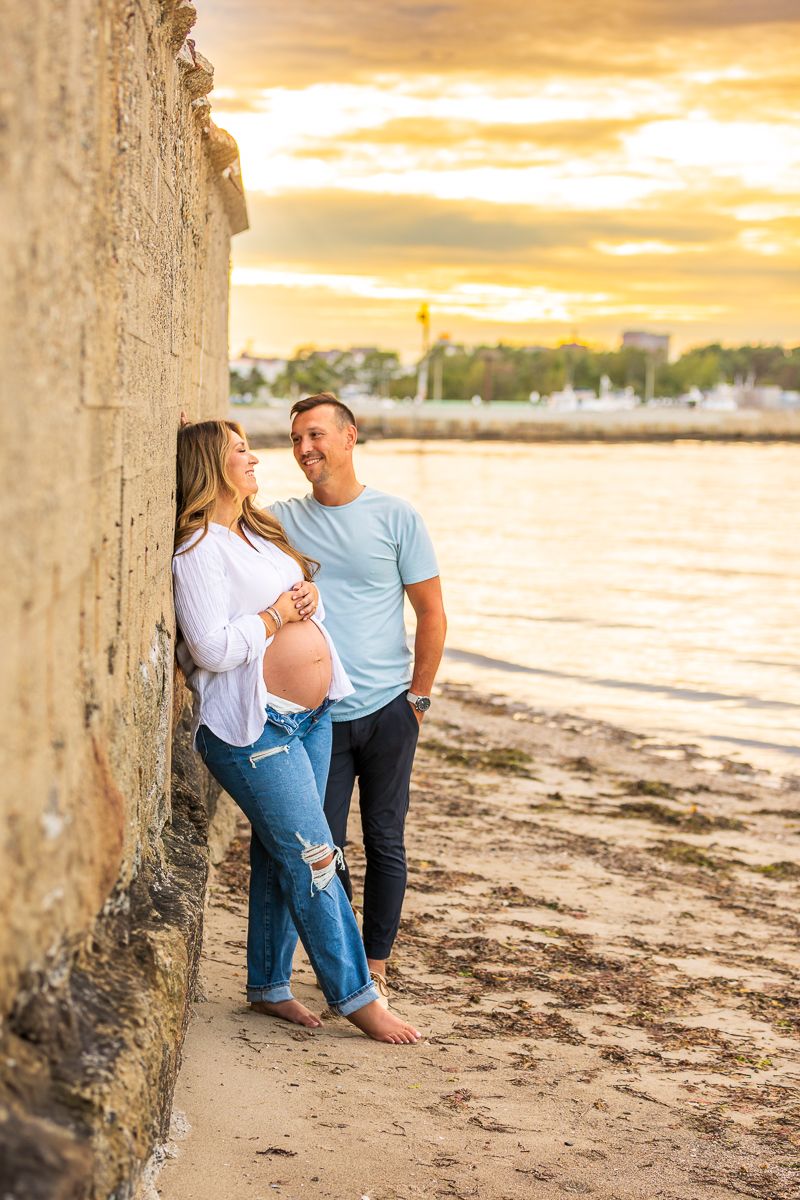 Expectant couple shares a tender moment at sunset on a beach near a concrete wall.