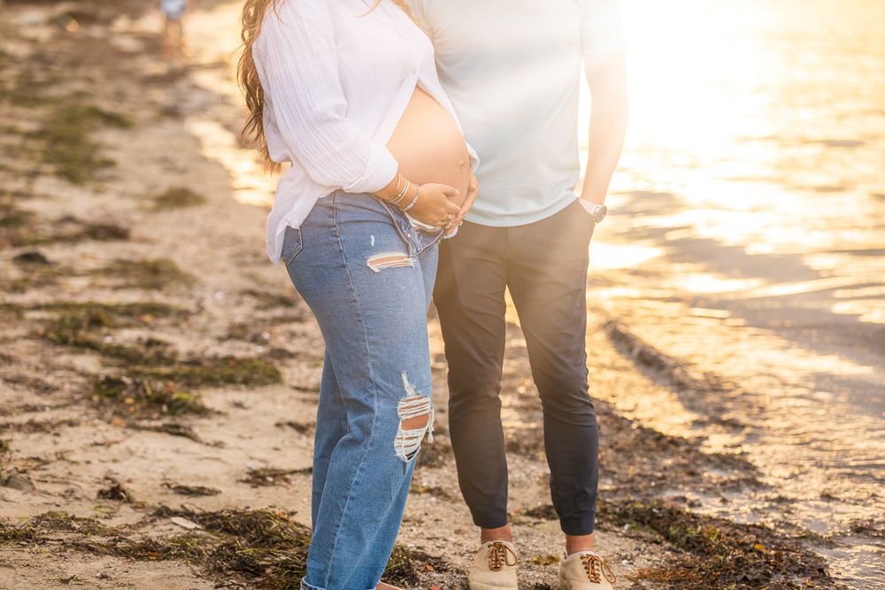 Couple standing on sandy beach during golden sunset holding hands while wearing casual denim and white clothing.