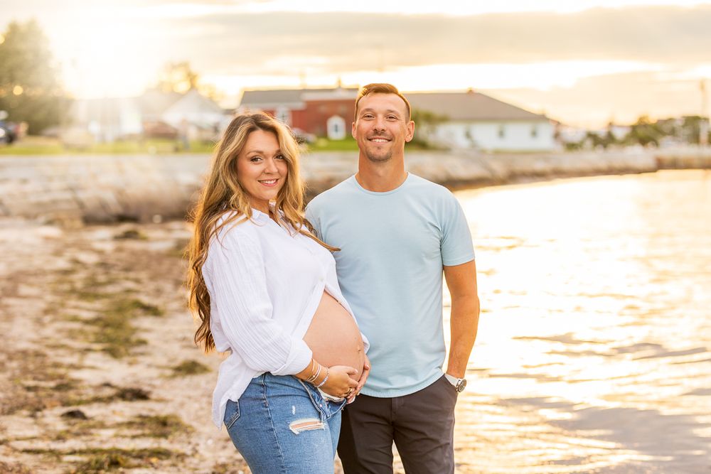 A couple stands together on a beach during golden hour sunset with water and houses in the background.
