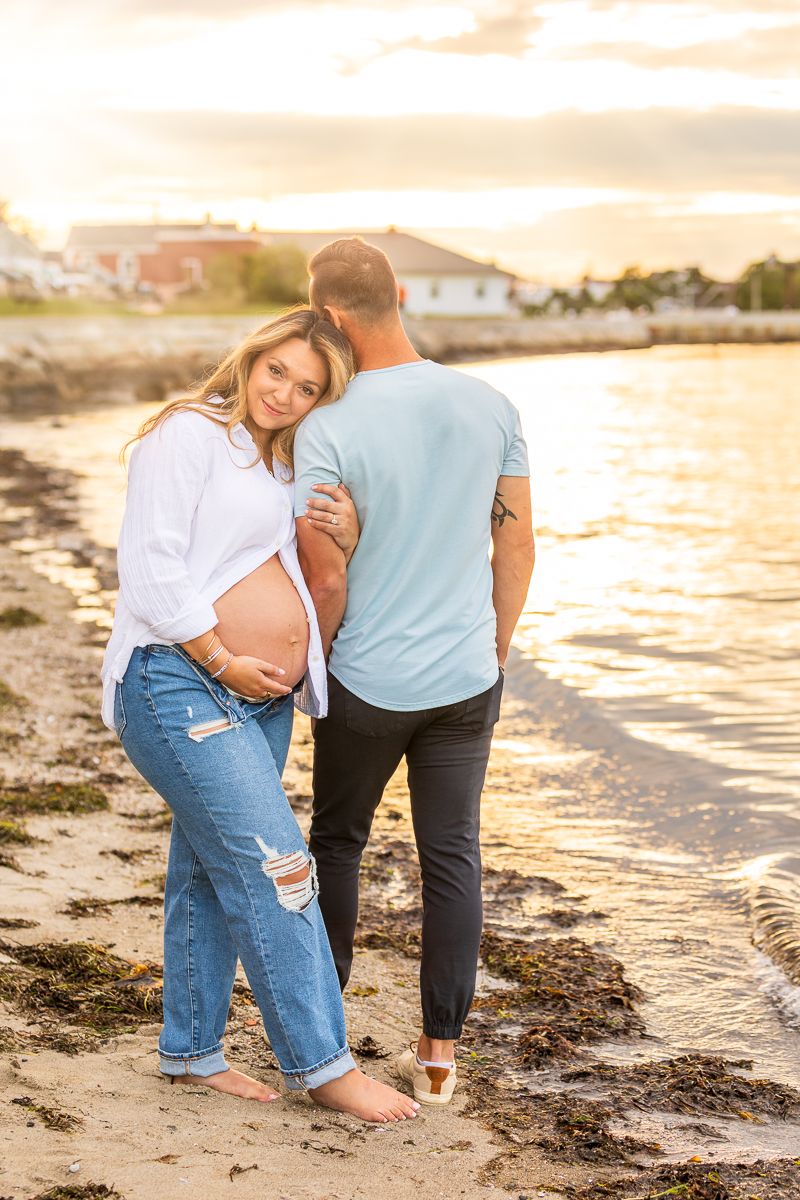 Couple shares intimate moment on beach at sunset while expecting a baby as waves lap at their bare feet.