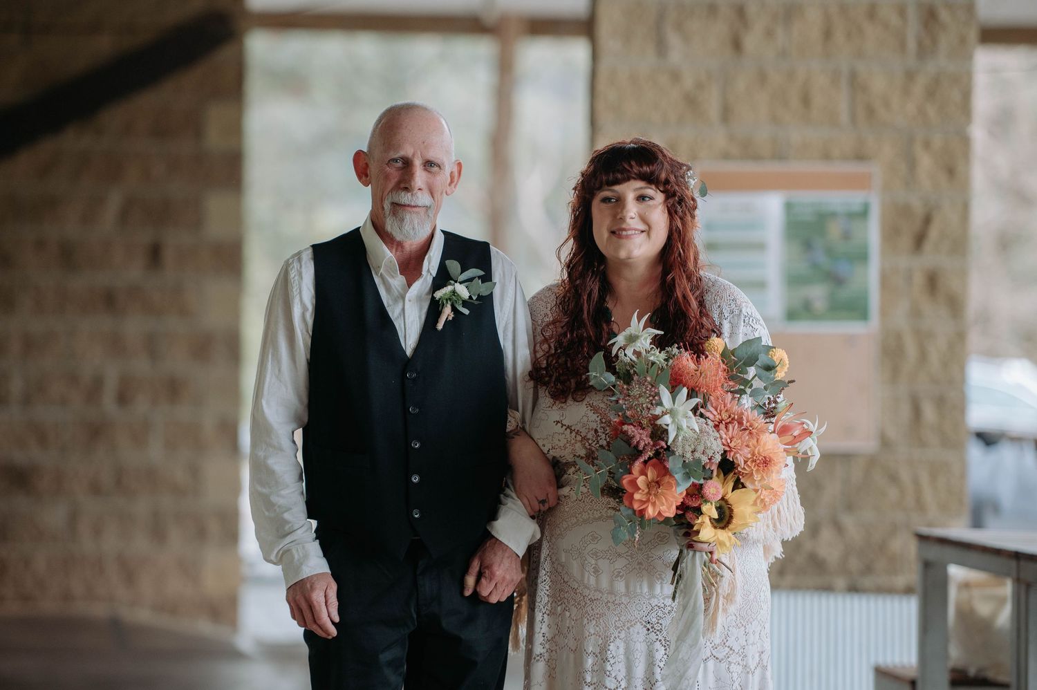 A bride and father figure stand together at a rustic stone venue, with the bride holding a colorful autumn bouquet.