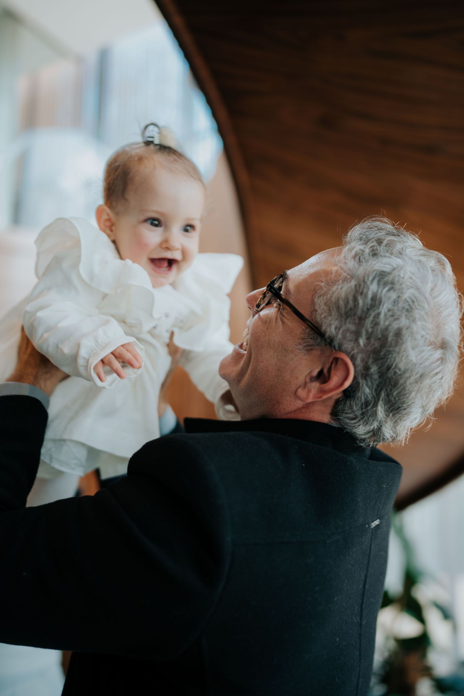 Baby in white dress being lifted up by older person in black sweater in heartwarming indoor moment.