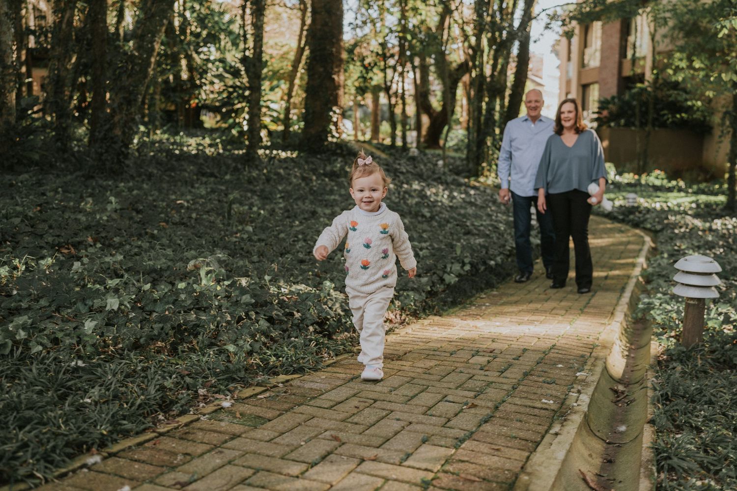Small child runs ahead on brick pathway while adults follow behind in wooded setting.