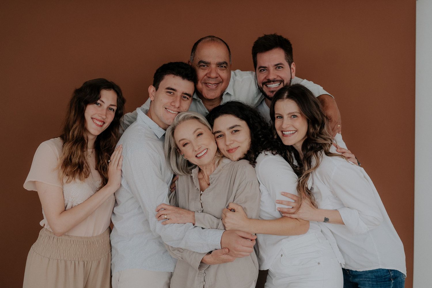 Large family group poses together in coordinated neutral outfits against brown background.