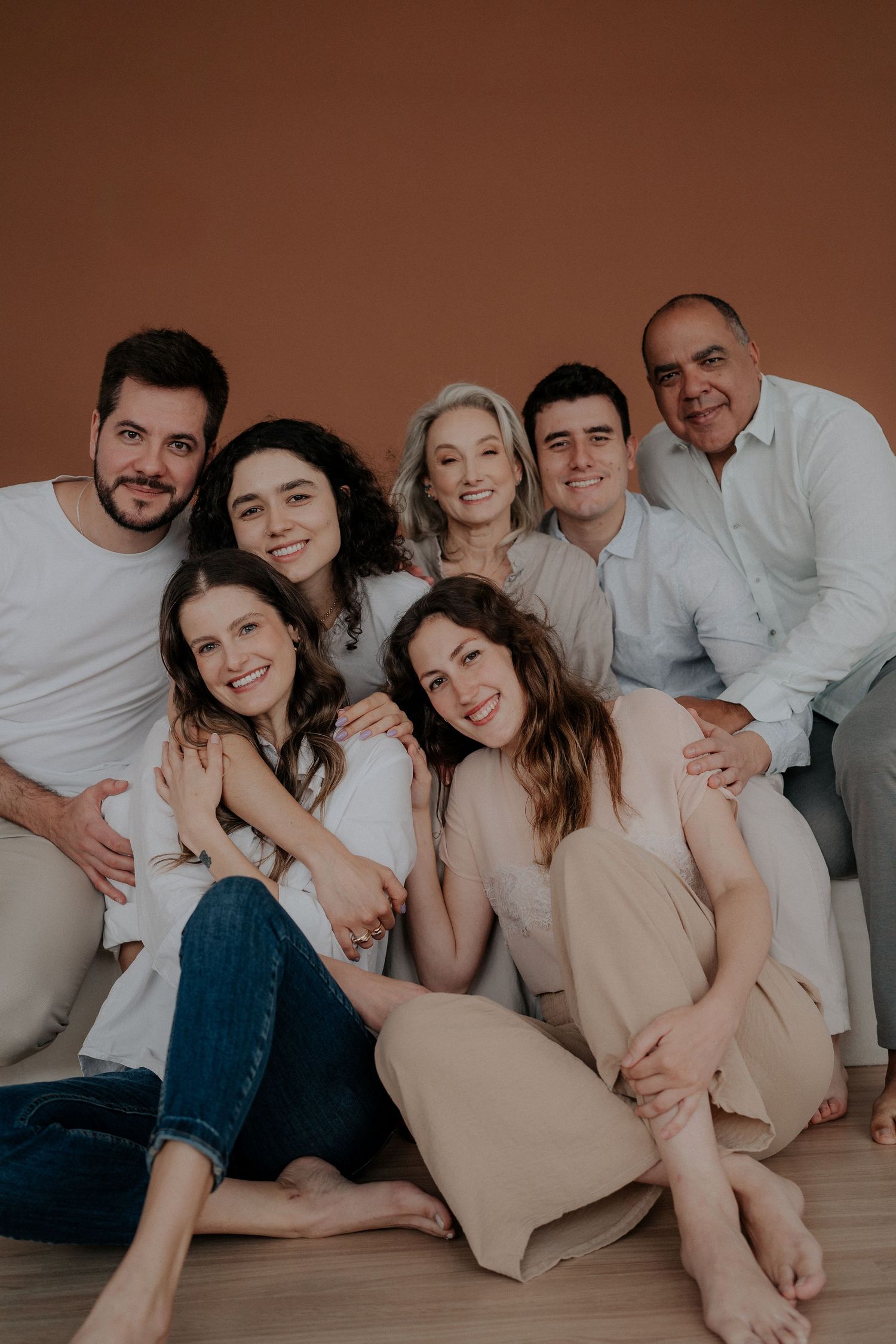 Family portrait session in casual neutral outfits lying close together against brown background.
