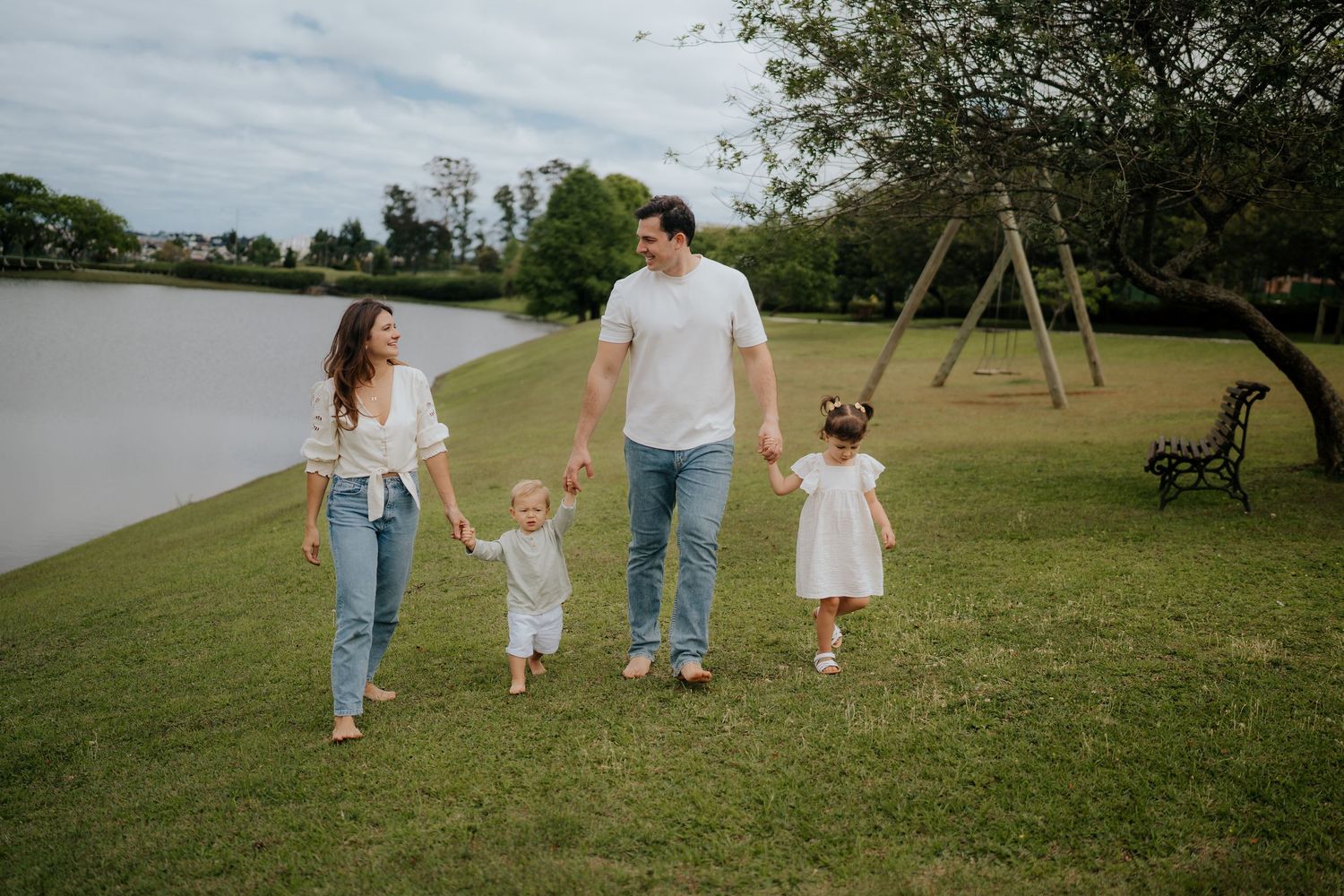 Family walking together on grassy lawn by a lake with playground swings in the background.