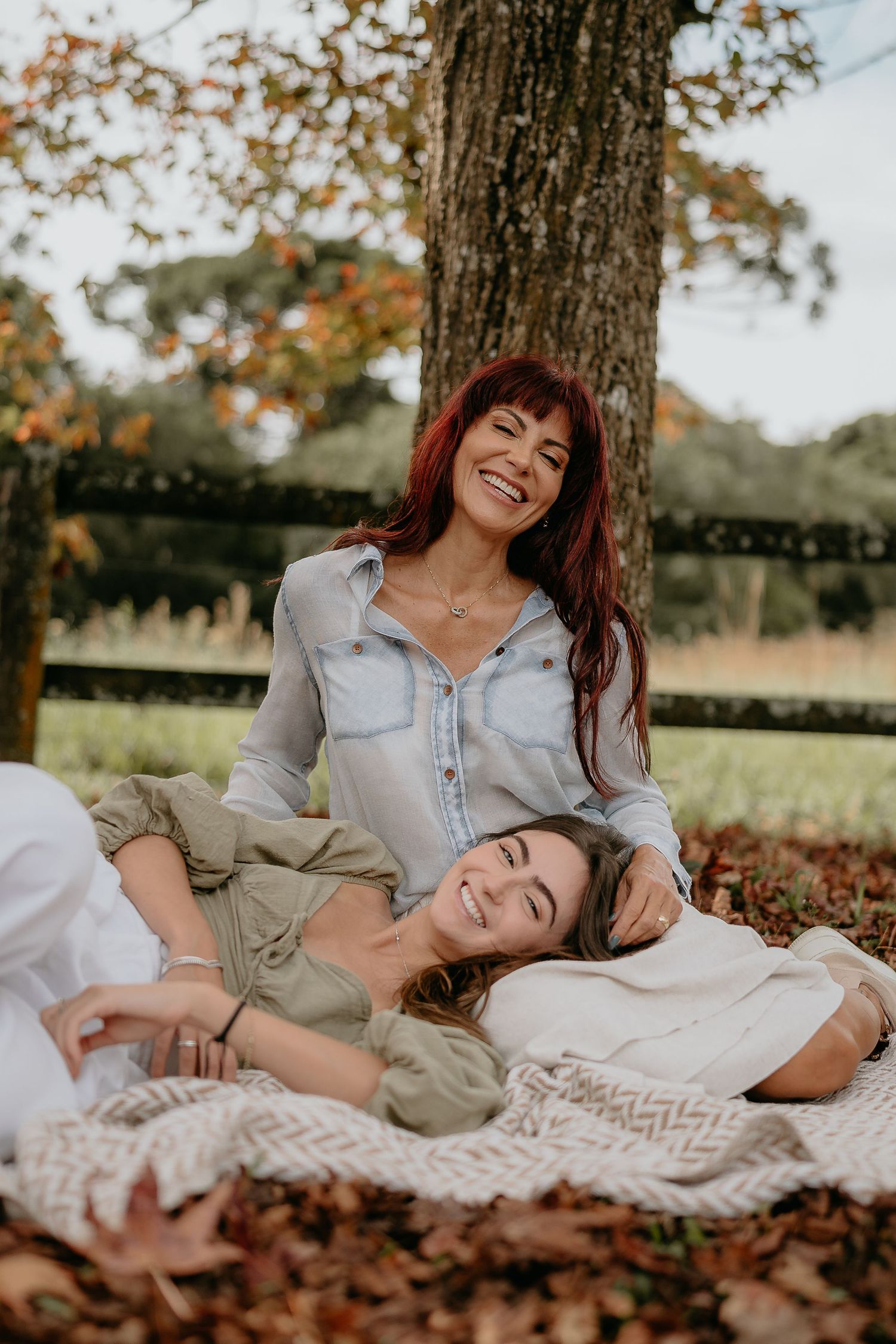Two friends sharing laughter while lying on a blanket under an autumn tree.