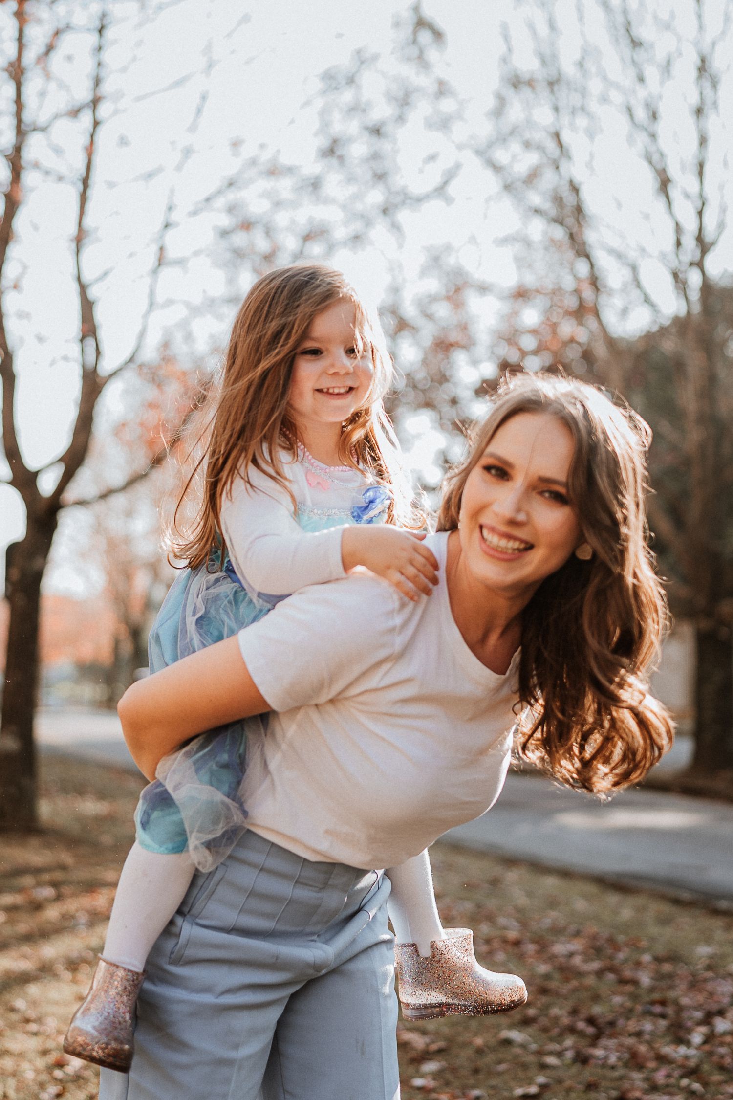 Mother giving daughter piggyback ride outdoors during fall season photoshoot.