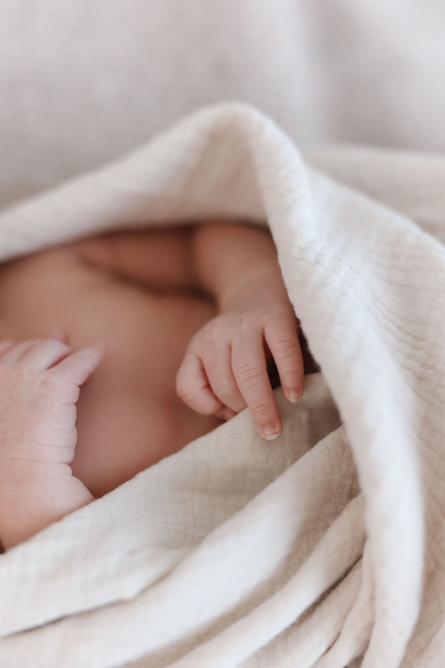 A tiny newborn baby hand reaches out from beneath a soft white blanket in a tender close-up photograph.